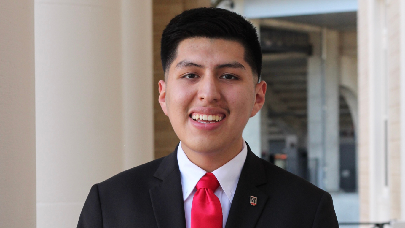A person wearing a black suit jacket with an arch pinned on the lapel, white dress shirt, and bright red tie stands outdoors near a columned building, posing.