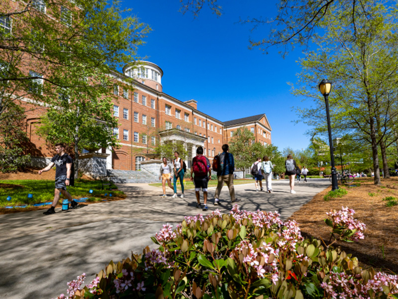Students walk along a sunny campus pathway lined with trees and spring flowers, with a large multi‑story brick academic building in the background under a bright blue sky.