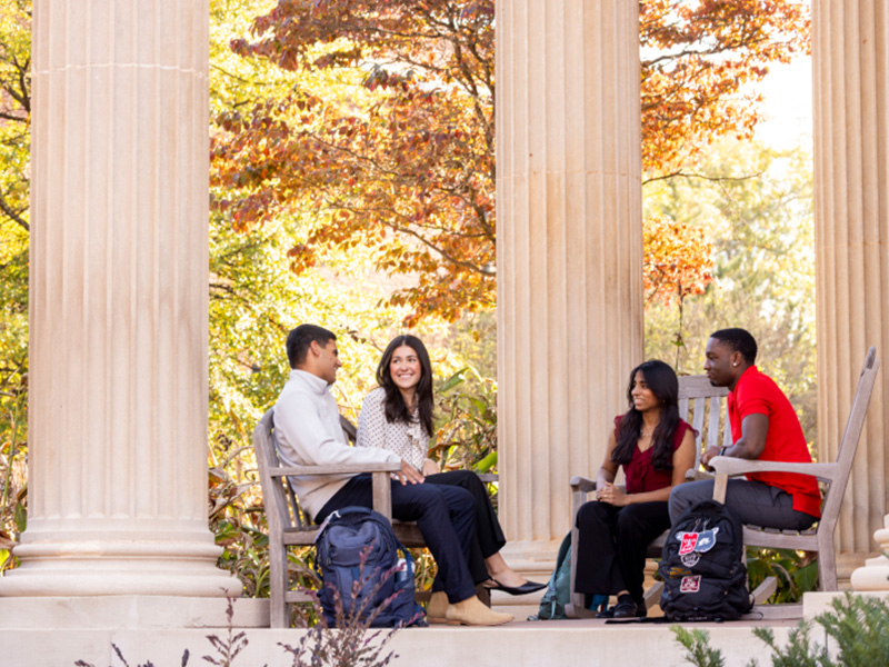 Four students sit in wooden chairs on a shaded outdoor terrace framed by large stone columns. They talk together with backpacks at their feet, surrounded by fall foliage and warm sunlight.