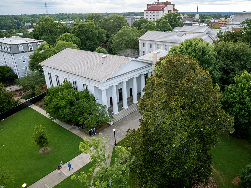 Aerial view of a historic white building with tall columns, surrounded by large trees and green lawns. A few people walk along the sidewalk below, and additional campus and city buildings are visible in the background.