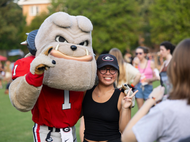 apply portal_2 A person poses and smiles for a photo with a bulldog mascot wearing a red jersey with the number 1. They are outdoors on grass, with trees and other people in the background. The person is making a peace sign.