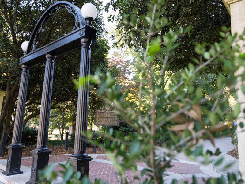 A view of a historic black iron arch with three tall columns and round white globes on top, standing along a brick pathway surrounded by dense green trees and shrubs. A historic marker sign is visible behind the arch, and sunlight filters softly through the foliage, creating a shaded, peaceful campus entrance scene.