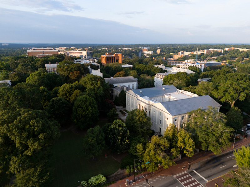 campus 4 An aerial view of a large, tree‑filled campus with academic buildings scattered among dense green foliage. A prominent light‑colored building with a gray roof sits near the foreground, bordered by a street and crosswalk. Additional campus structures extend into the distance, including several mid‑rise brick buildings. The horizon shows a wide expanse of forested landscape under a soft, partly cloudy sky.