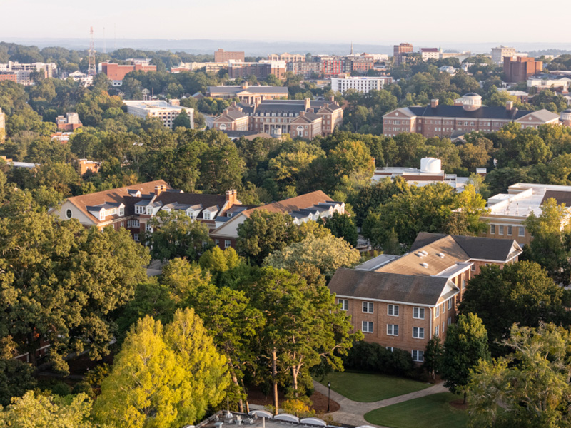 An aerial view of a tree‑filled campus with a mix of academic buildings and residential structures surrounded by dense greenery. Light-colored and red‑brick buildings are scattered throughout the landscape, with sidewalks and small roads weaving between them. In the distance, additional campus facilities and city buildings extend toward the horizon under soft daylight.