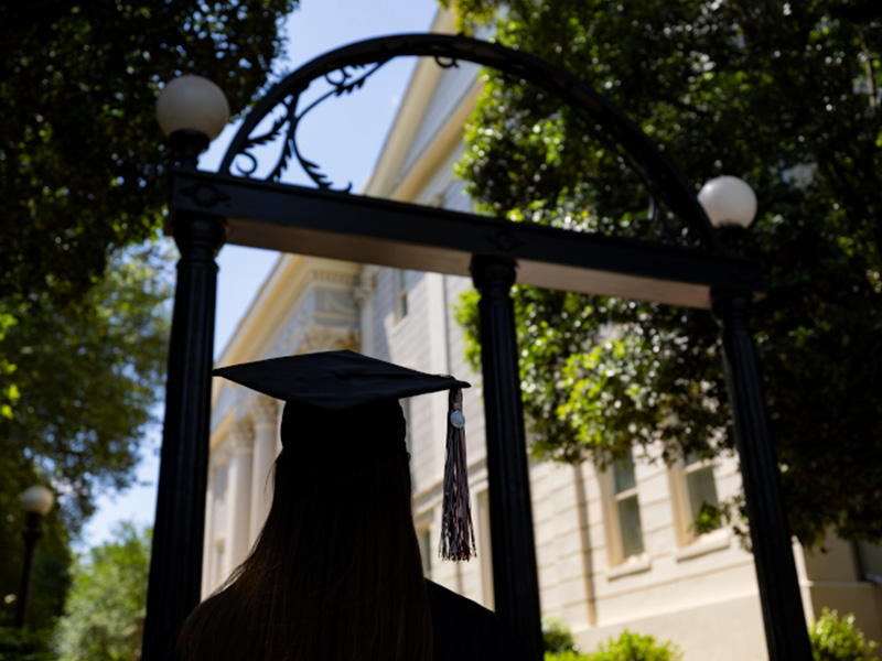 A graduate wearing a cap and gown stands facing a historic black iron arch, framed by tall trees and bright sunlight. The tassel hangs from the graduation cap, and a light-colored academic building is visible in the background through the archway. The scene captures a symbolic moment of reflection and transition on campus.