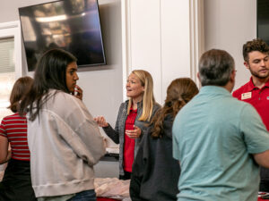 A group of people stand in a room having a conversation near a table covered with materials. One person wearing a red shirt and name tag speaks with attendees, while others gather around and listen. A large wall-mounted TV is visible in the background.