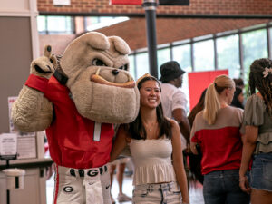 A costumed bulldog mascot wearing a red football jersey poses with a person in a sleeveless white top inside a busy indoor event space, with other attendees gathered and walking in the background.