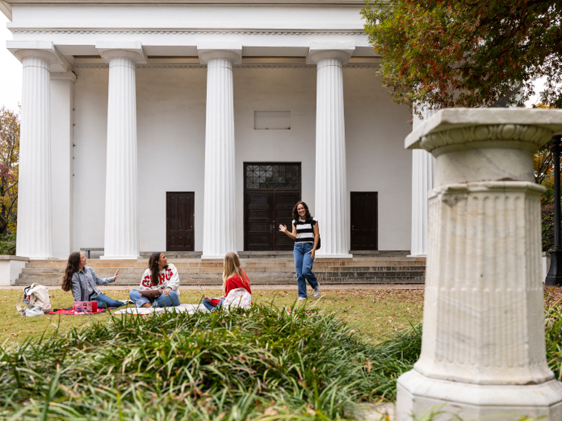 A group sits on a picnic blanket on a grassy lawn in front of a large white building with tall, fluted columns. One person stands and gestures toward the group, while others sit and talk. A stone pedestal is visible in the foreground, and colorful autumn foliage frames the scene.