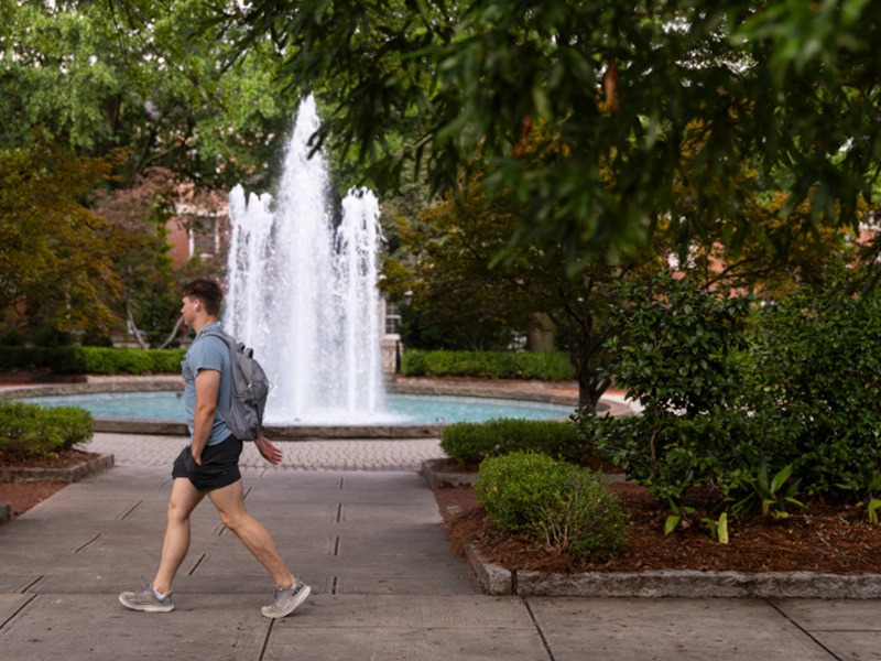 A person walks along a paved campus pathway lined with neatly trimmed shrubs and trees, passing in front of a large fountain with tall, white jets of water. The surrounding greenery frames the scene, and the fountain sits at the center of a landscaped circular pool. The setting appears calm and shaded, with soft light filtering through the leaves.