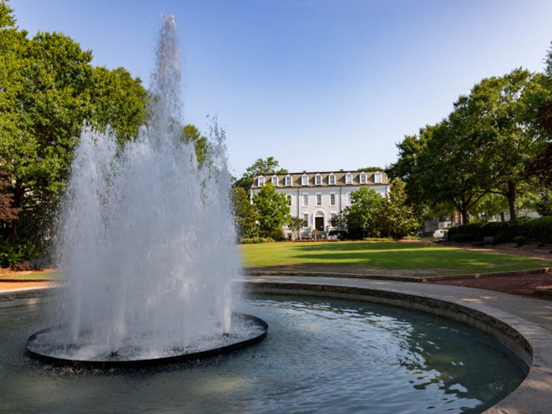 herty and honors A large water fountain in the foreground with a circular basin, surrounded by green lawns and trees, with a multi-story white academic building in the background under a clear blue sky.