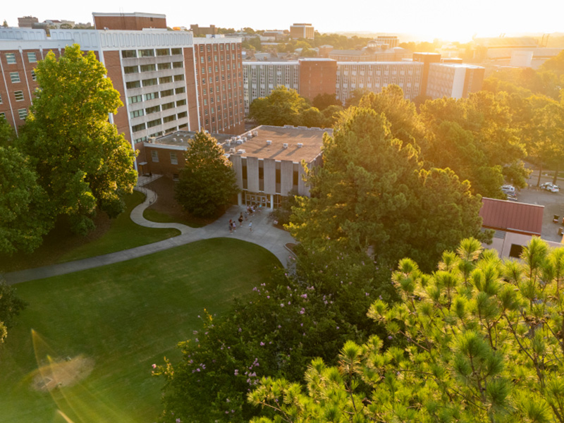 move-in An aerial view of a university campus at sunset, showing large residence hall buildings surrounded by trees and green lawn areas, with warm sunlight casting a golden glow across the scene.