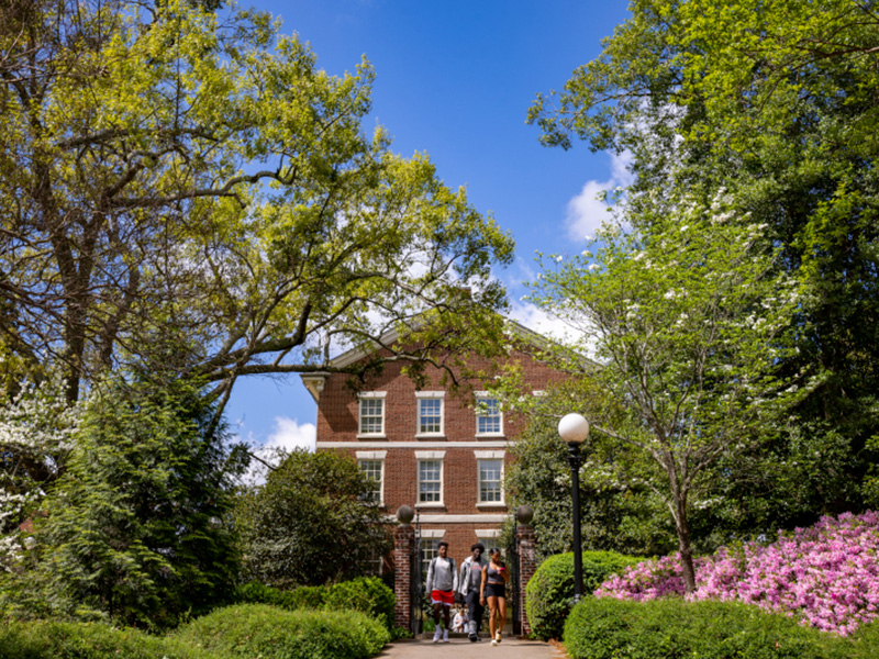 A group of people stands on a path in front of a red-brick building surrounded by lush greenery, blooming flowers, and tall trees under a bright blue sky.