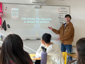 A student stands at the front of a classroom giving a presentation titled “The Road to College & What to Expect” projected onto a whiteboard. Several classmates sit at desks facing the presenter, and handwritten notes are visible on a whiteboard to the side.