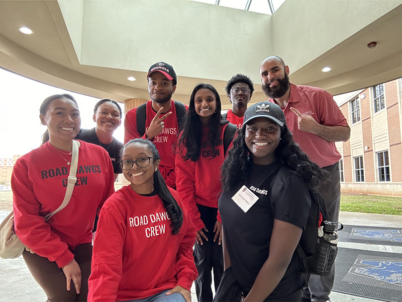 A group of people wearing matching “Road Dawgs Crew” shirts pose together in a bright indoor space with large windows overhead. Most of the group is dressed in red crew shirts, while one person in the front wears a black version with a name tag. They are standing and crouching closely for a group photo, with a brick building visible through the windows behind them.