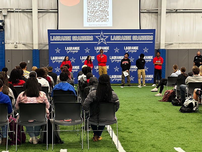 An audience seated on chairs in an indoor athletic facility listens to a group of speakers standing at the front of the room. The speakers stand in front of a blue backdrop with repeated text reading “LaGrange Grangers” and star logos. A large screen above the backdrop displays a QR code. The setting appears to be a school or community event held on artificial turf inside a large gym-like space.