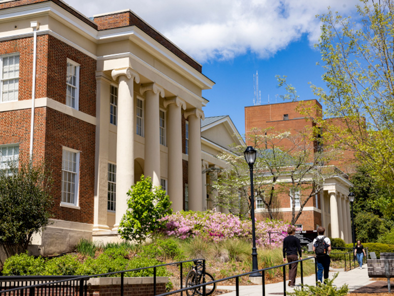 spring A red‑brick academic building with tall white columns stands along a landscaped campus walkway. Blooming pink flowers and green shrubs line the path in front of the building, and trees with spring foliage frame the scene. A few people walk along the paved path under a bright sky with patches of clouds.