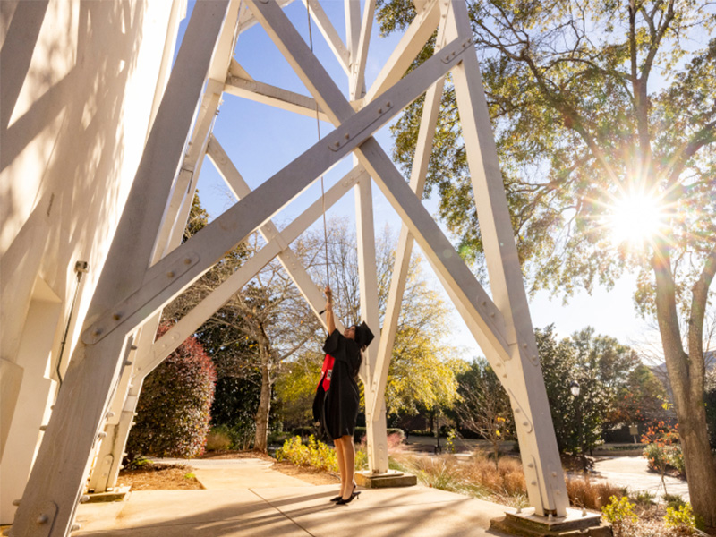 student ringing bell A graduate wearing a cap and gown rings the bell beneath a large white wooden bell tower structure on a sunny day. Sunlight streams through the trees, casting long shadows across the walkway and surrounding landscaping. The scene captures a celebratory campus tradition framed by bright blue sky and greenery.