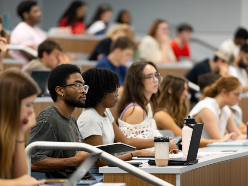 A group of college students sit in a lecture hall, attentively listening and taking notes. Laptops, notebooks, and drinks are on their desks. The room is brightly lit and has tiered seating.