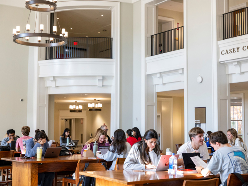 students studying Students study and work at large wooden tables inside a bright, spacious academic building with high ceilings and white architectural columns. Laptops, notebooks, and water bottles are spread across the tables as students read, type, and collaborate. Large chandeliers hang from the ceiling, and upper-level balconies overlook the study area. A sign reading “Casey Commons” is visible on the right above an entryway.