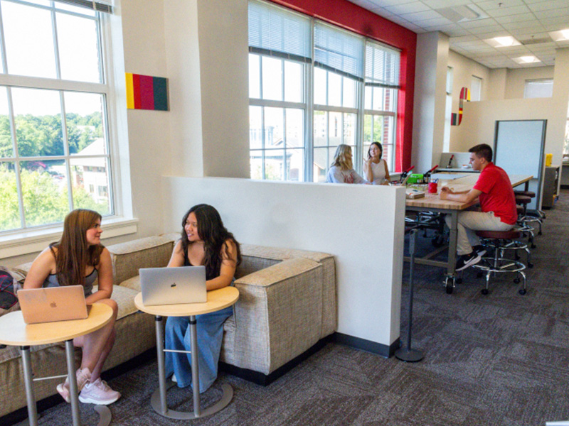Students work and study inside a bright, modern campus space with large windows overlooking trees. Two students sit on a cushioned corner booth with laptops on small round tables, while others gather at a high table in the background. The room features light-colored walls, red accents, and soft natural light filling the space, creating an open and collaborative study environment.