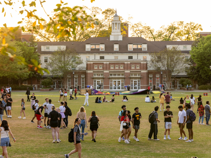 Students gather and walk across a large campus lawn at sunset, with groups talking, sitting, and relaxing in front of a multi‑story brick academic building. Trees frame the scene as warm light falls across the open green space.
