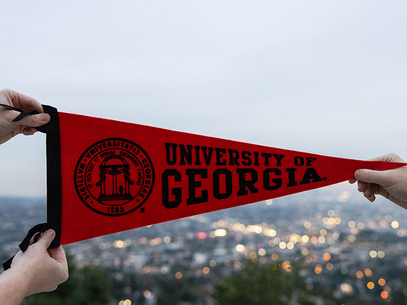 Two hands hold up a red pennant that reads “University of Georgia” with the UGA Arch seal printed on the left side. The scene is set outdoors at dusk, with a cityscape of blurred lights and buildings in the background under a soft, overcast sky.