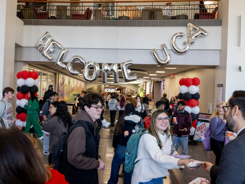 A busy campus hallway decorated with large silver balloon letters spelling “WELCOME UGA,” along with red, black, and white balloon columns. Students walk through the space and interact at tables as part of a welcome event.