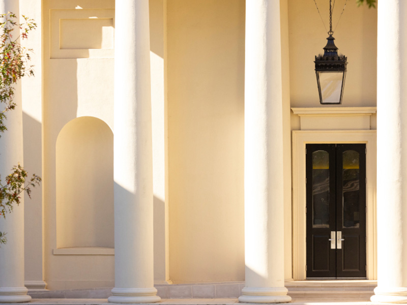 white building The exterior of a light‑colored building with tall, smooth columns framing a black double door. A decorative hanging lantern is suspended above the entrance, and soft sunlight creates gentle shadows across the façade. Architectural details, including an arched inset niche and clean geometric lines, add to the classical, elegant appearance of the structure.